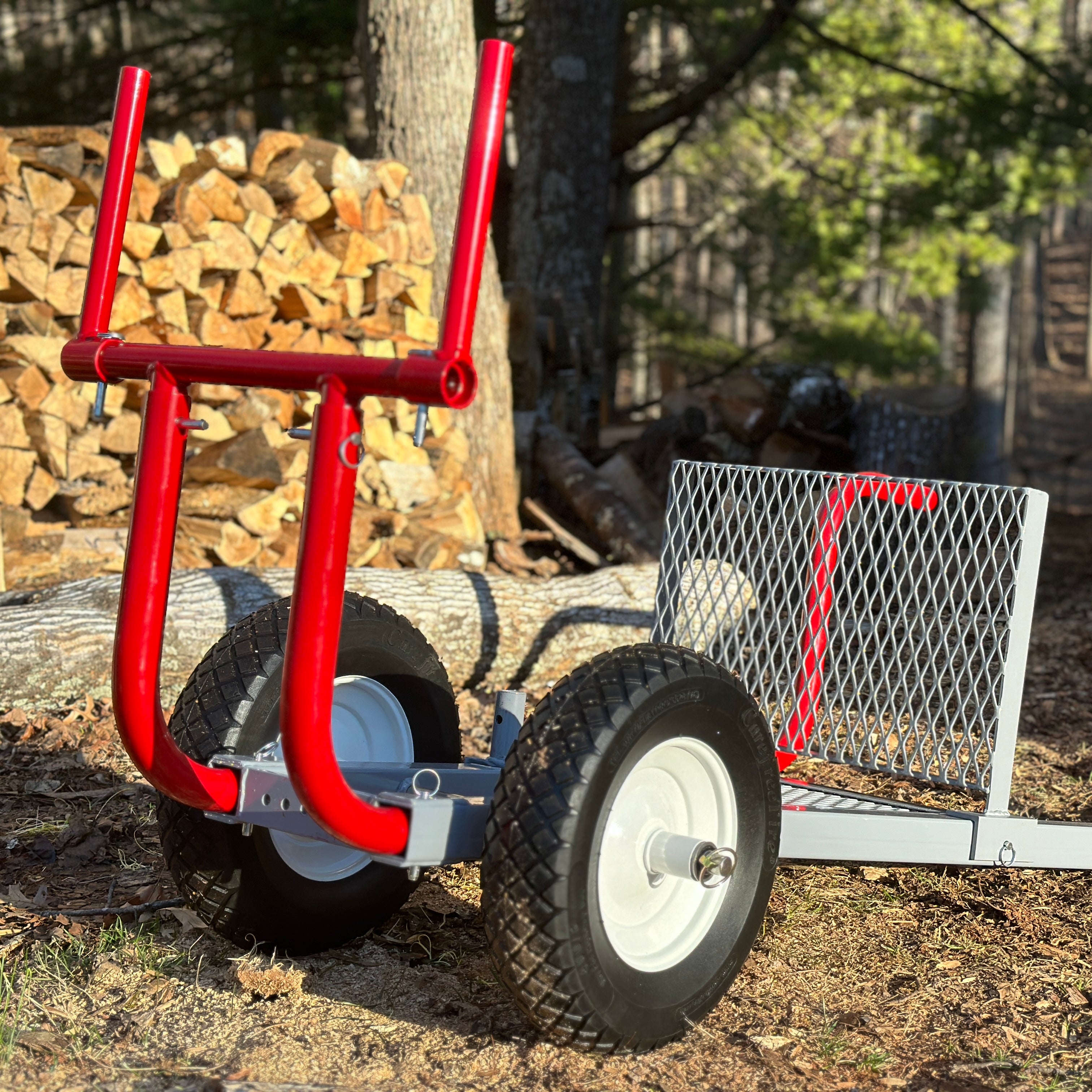 Red metal log carrier with wheels on a forest floor with stacked logs in the background.