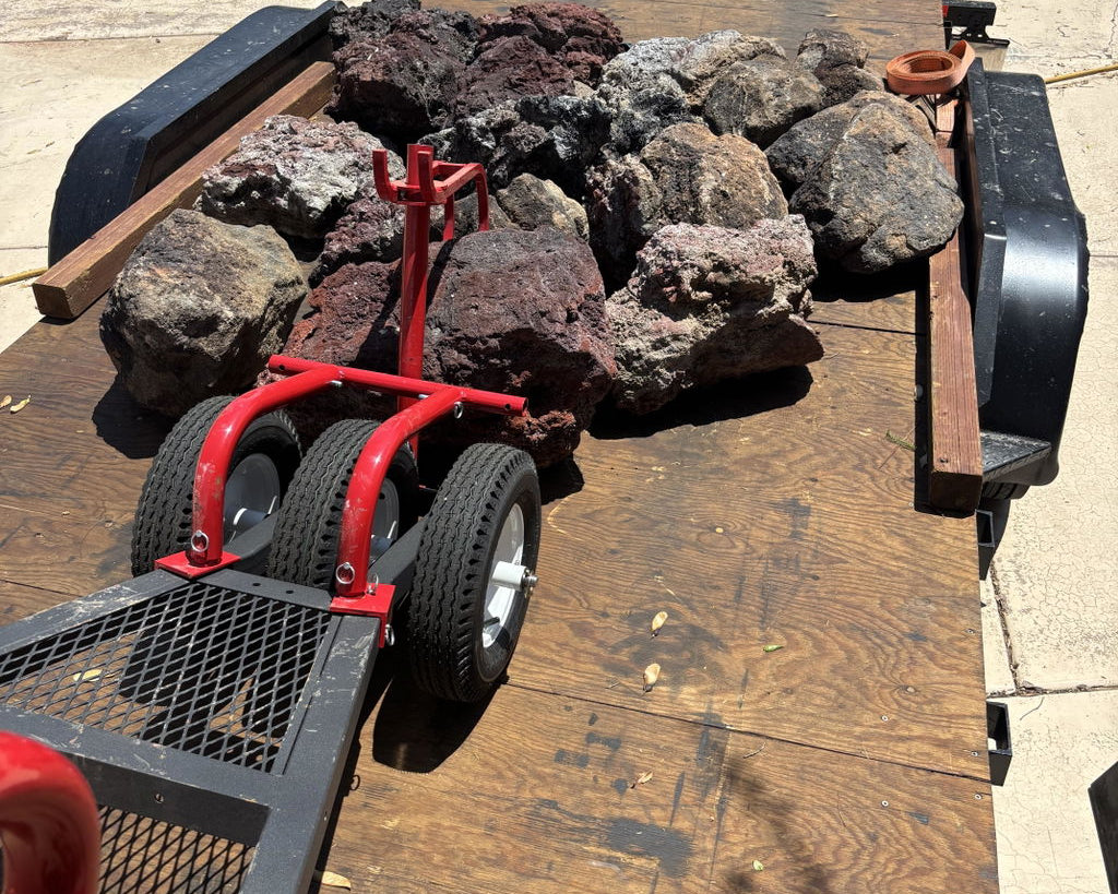 A red metal rock spreader and trailer jack positioned on the tongue of a wooden flatbed trailer loaded with large decorative river rocks, photographed outdoors on a concrete surface.