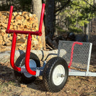 Red metal log carrier with wheels on a forest floor with stacked logs in the background.