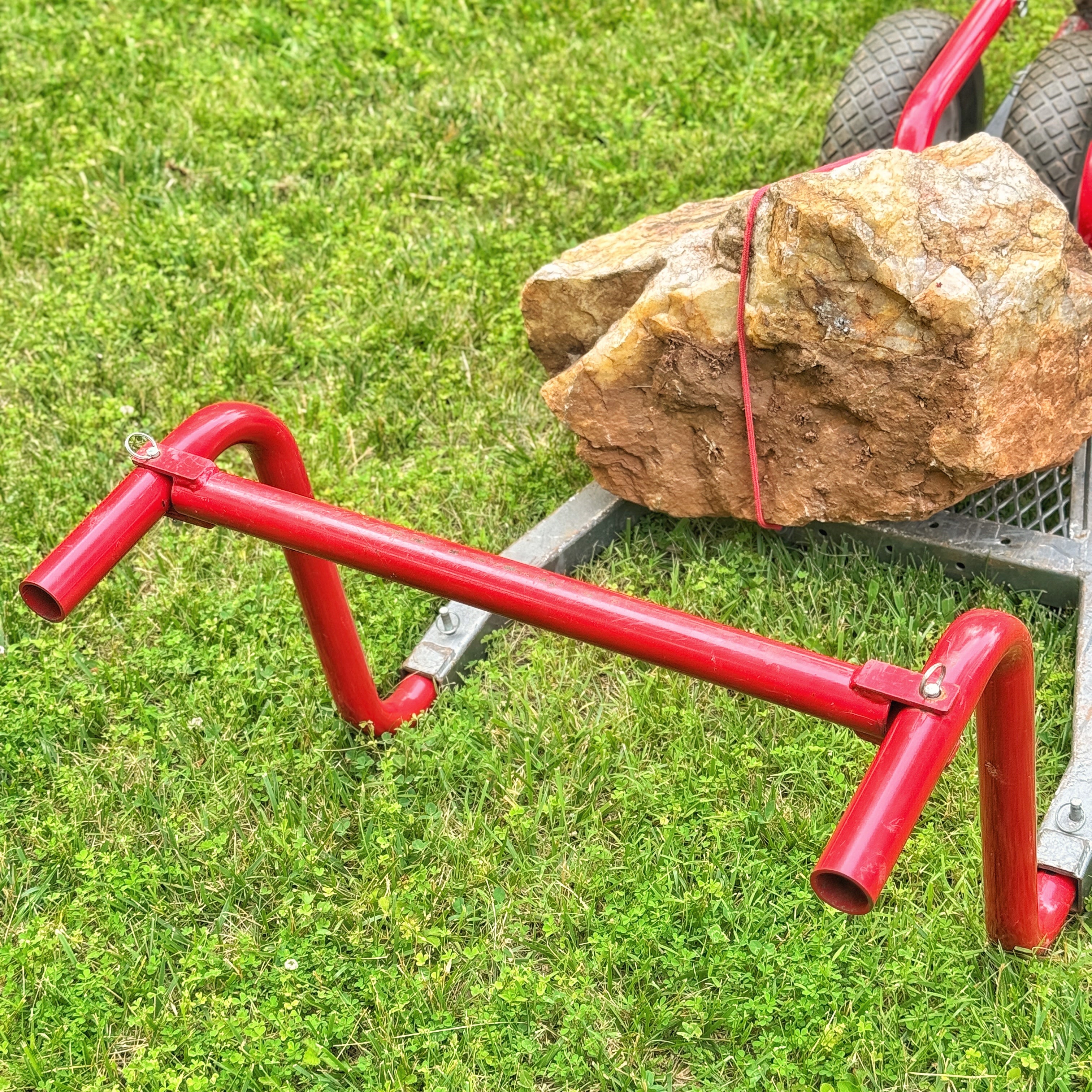 Red metal bar attached to a cart with a large rock on grass