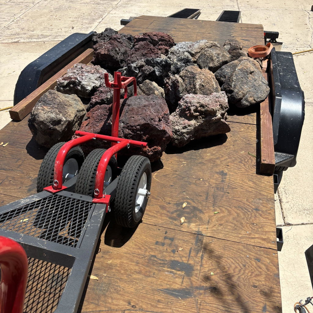 A red metal rock spreader and trailer jack positioned on the tongue of a wooden flatbed trailer loaded with large decorative river rocks, photographed outdoors on a concrete surface.