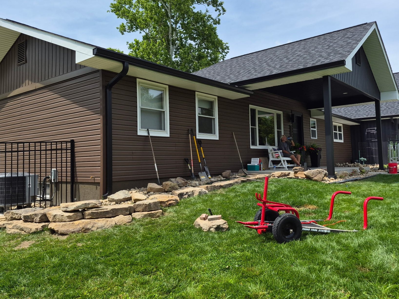 a brown house with a stone retaining wall and a rock moving cart in the front yard